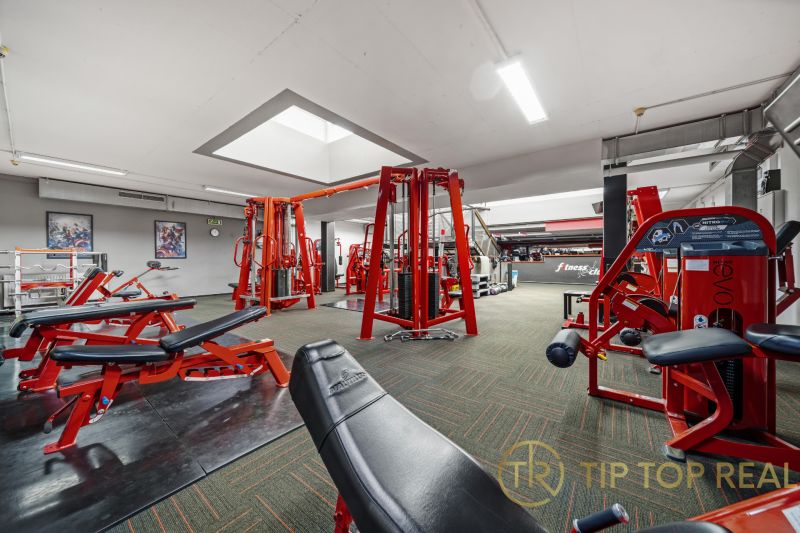 Fitness centrum s posilovacími stroji a červenými lavičkami, vhodné obchodní prostory.