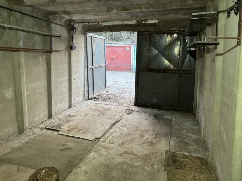 Betonová garáž s otevřenými ocelovými dveřmi, pohled na další garáž venku.