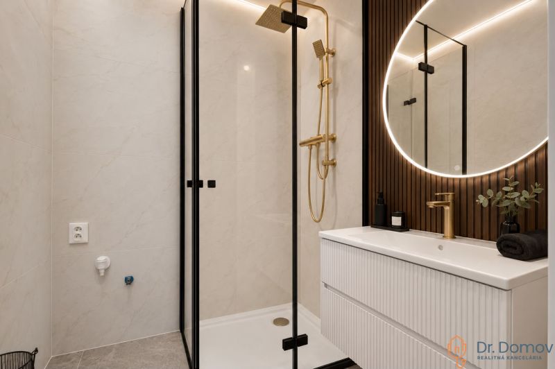 Koupelna ve 3-pokojovém bytě s elegantní sprchou, zlatými detaily a osvětleným zrcadlem.