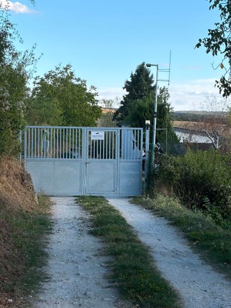 Brána vedoucí k chatě v Bance obklopená zelení a polní cestou.