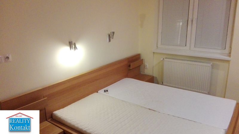 Ložnice třípokojového bytu s dřevěnou postelí a oknem s roletami na stěně.