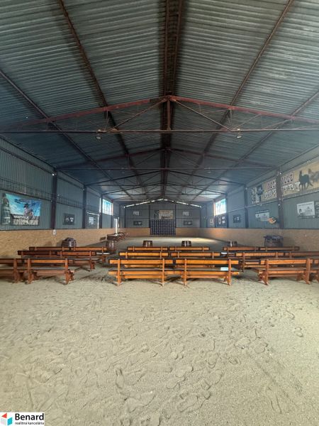 Interiér budovy s dřevěnými lavicemi a písčitou podlahou, určený pro akce.