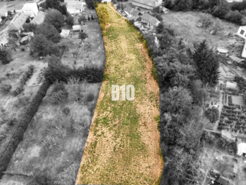 Pozemky - bydlení Kolíňany: Dlouhý pozemek se zelenou trávou, lemovaný stromy a stavbami.