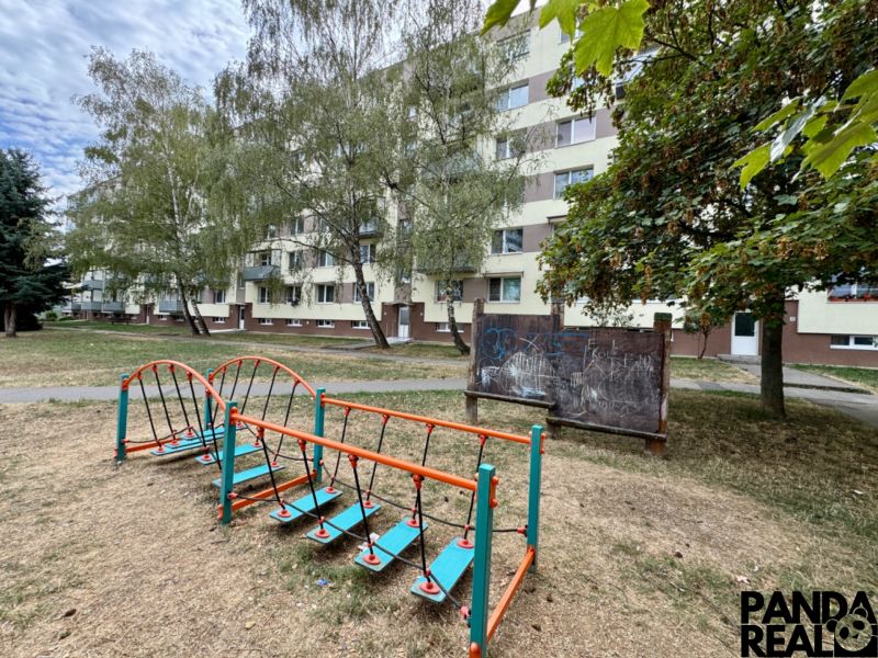 Exteriér bytovky v Topoľčanech s dětským hřištěm a vegetací kolem 1-pokojového bytu.