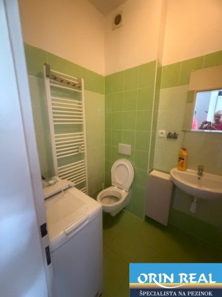 Koupelna garsonky se zeleným obkladem, umyvadlem, toaletou a pračkou.