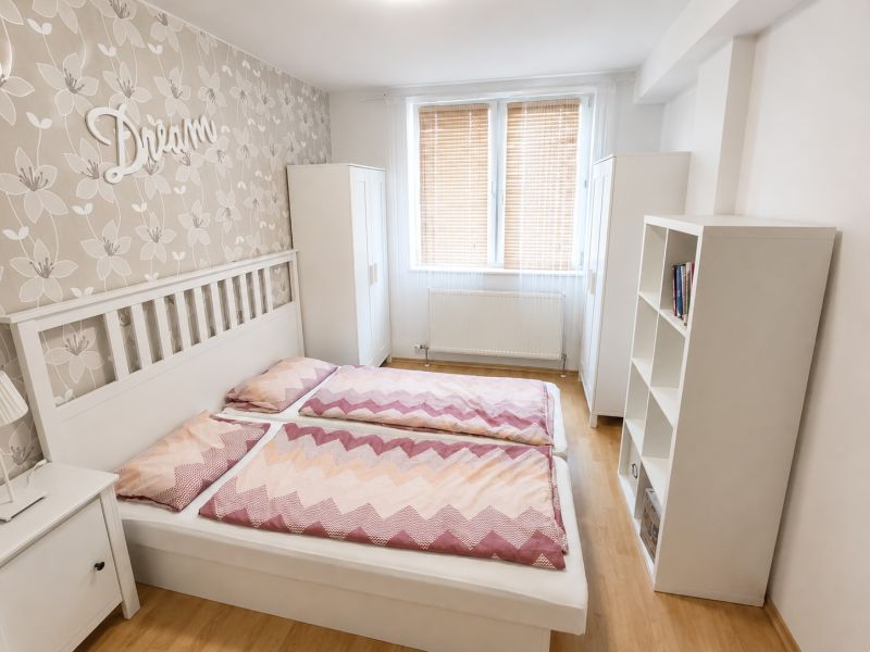 Světlá ložnice s manželskou postelí, květinovou tapetou a podlahou s dřevěným dekorem.