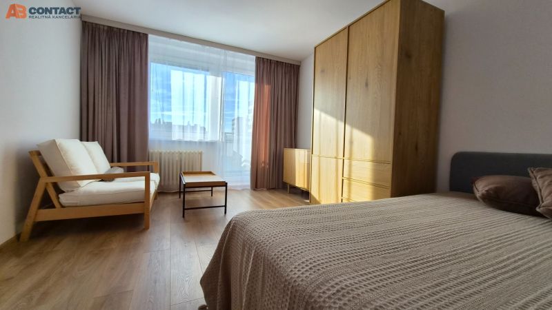 Pokoj s velkým oknem, gaučem, postelí, skříní a podlahou s dřevěným dekorem v 1-pokojovém bytě.