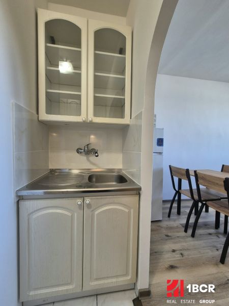 Kuchyňský kout v garsonce s dřezem, skříněmi a stolem na podlaze s dřevěným dekorem.