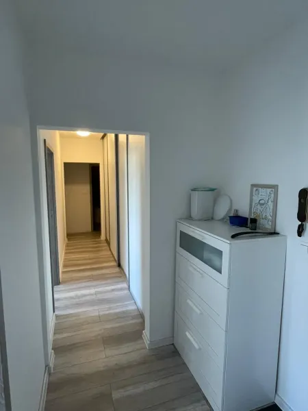 Chodba v 2-pokojovém bytě s bílou komodou a podlahou s dřevěným dekorem.