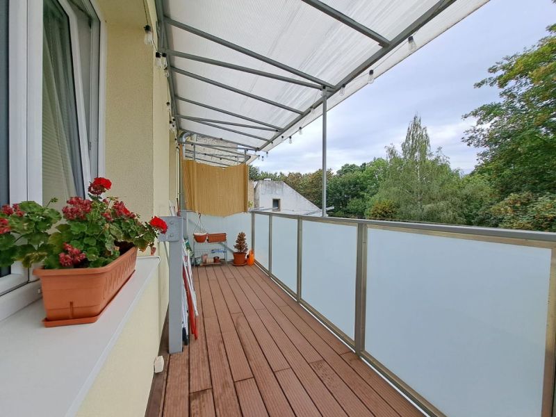 Balkon v 3-pokojovém bytě s podlahou s dřevěným dekorem, květinami a výhledem.