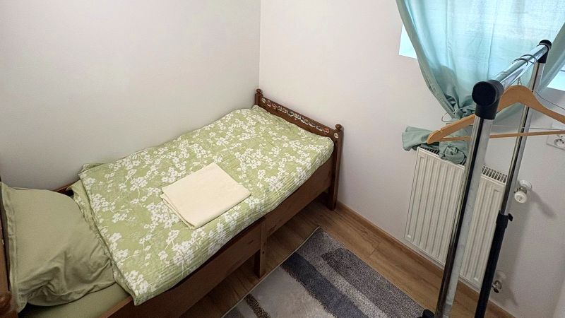 Jednolůžková postel, stojan na oblečení, zelené závěsy, podlaha s dřevěným dekorem.