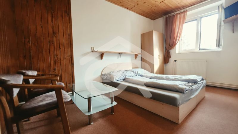 Ložnice rekreačního apartmánu s manželskou postelí, křesly a oknem, podlaha s dřevěným dekorem.