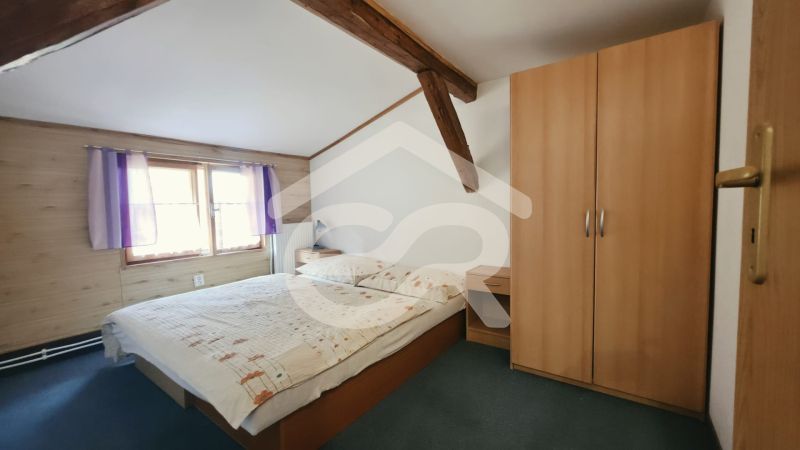 Ložnice v rekreačním apartmánu s dřevěnými trámy, skříní a postelí vedle okna.