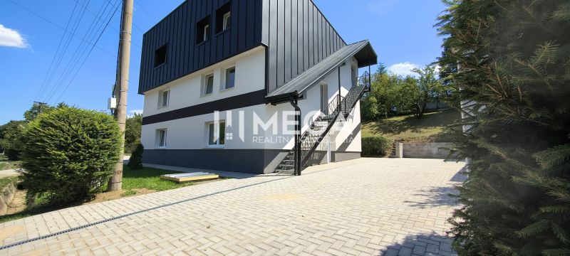 Čtyřpatrová budova v Jasenově s venkovními schody, obklopená zelení a dlážděným chodníkem.