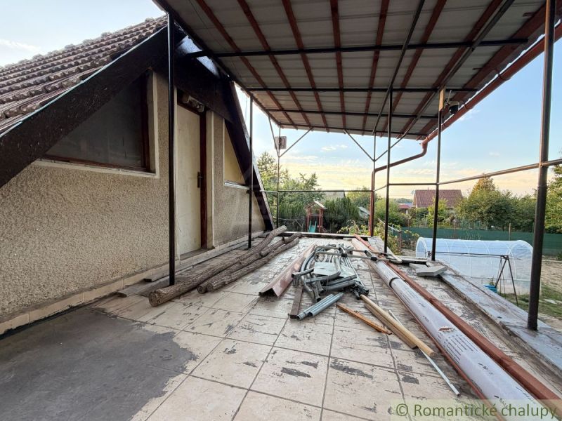 Terasa chaty v Nových Zámcích s nepořádkem a stavebním materiálem, klidná zahrada v pozadí.