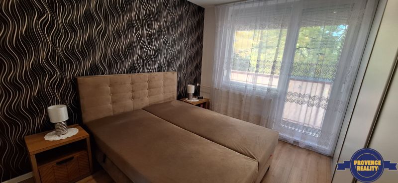 Ložnice s čalouněnou postelí, vzorovaná tapeta, záclony, podlaha s dřevěným dekorem.