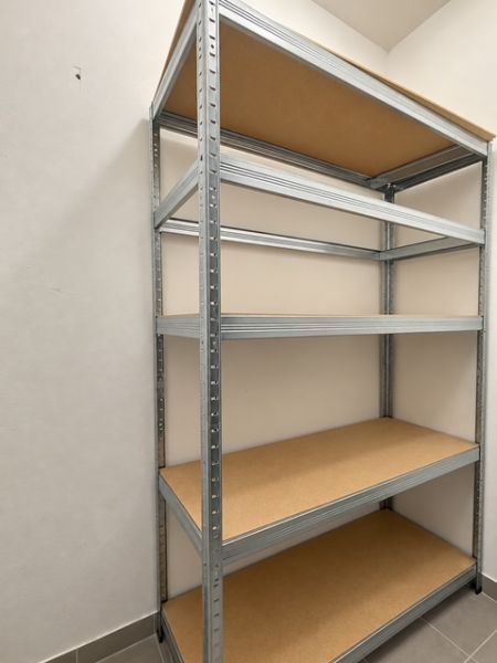 Kovový regál s policemi ve 2-pokojovém bytě na bílé stěně a šedé podlaze.