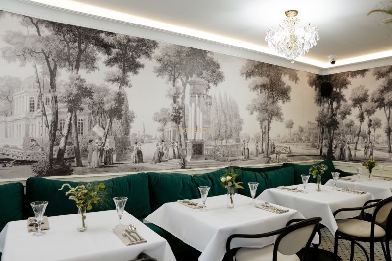 Restaurace s historickou tapetou, křišťálovým lustrem a zelenými sedačkami v obchodních prostorách.